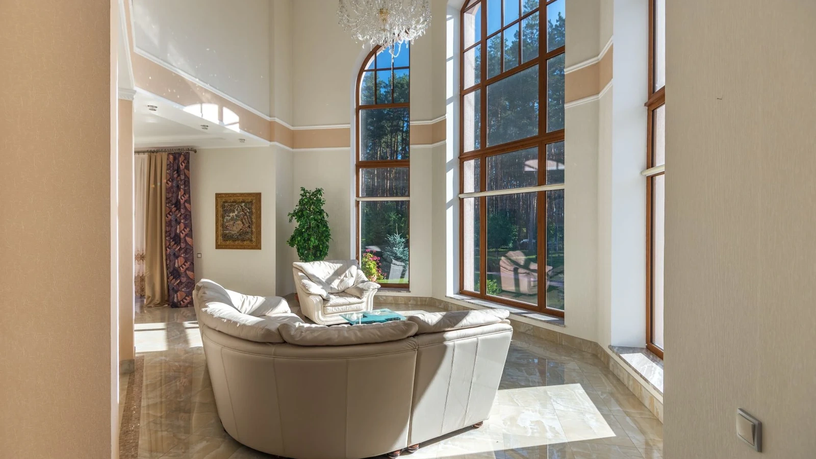 Tall arched wood-tone window frames and broad glass panes overlooking trees in a sunlit double-height living room.